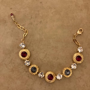 May include: A gold-toned bracelet featuring a series of round, textured settings. Each setting holds a gemstone in shades of red, clear, and gray. The bracelet has a chain closure and is displayed on a neutral background.