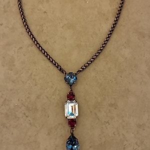 May include: A necklace featuring a braided copper-toned chain and a central pendant. The pendant consists of a blue crystal, two small red circular crystals, a large rectangular clear crystal, and a blue teardrop-shaped crystal at the base.