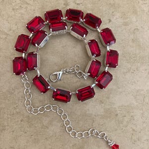 Op de afbeelding: Een ketting met grote, rechthoekige, robijnrode kristallen gezet in zilverkleurig metaal. De ketting heeft een ketting met een karabijnsluiting en een verlengketting met een kleine rode kristallen hanger.