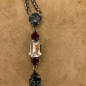 May include: A silver necklace with a series of colourful gemstones. The necklace features a large, rectangular clear gem, with smaller red and blue gems. The chain is a braided design.