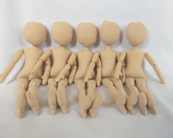 May include: Five unpainted fabric dolls sitting in a row. The dolls are beige and have simple features. They are all wearing the same outfit, which is a simple, beige dress.
