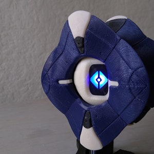 May include: A 3D-printed Ghost shell in blue and white, from the game Destiny. The shell has a glowing blue light in the centre and is displayed on a stand. The design features a geometric shape.
