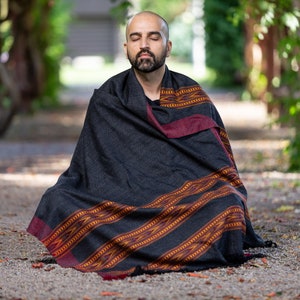 Pode incluir: Um homem está sentado de pernas cruzadas em um caminho de cascalho, meditando com os olhos fechados. Ele está vestindo um xale estampado em preto e vermelho com uma borda bordô.