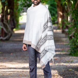 Puede incluir: Un hombre con un chal blanco con un patr&oacute;n geom&eacute;trico negro y marr&oacute;n. El chal tiene flecos en el borde inferior. El hombre lleva pantalones azules y est&aacute; de pie en un camino de grava.