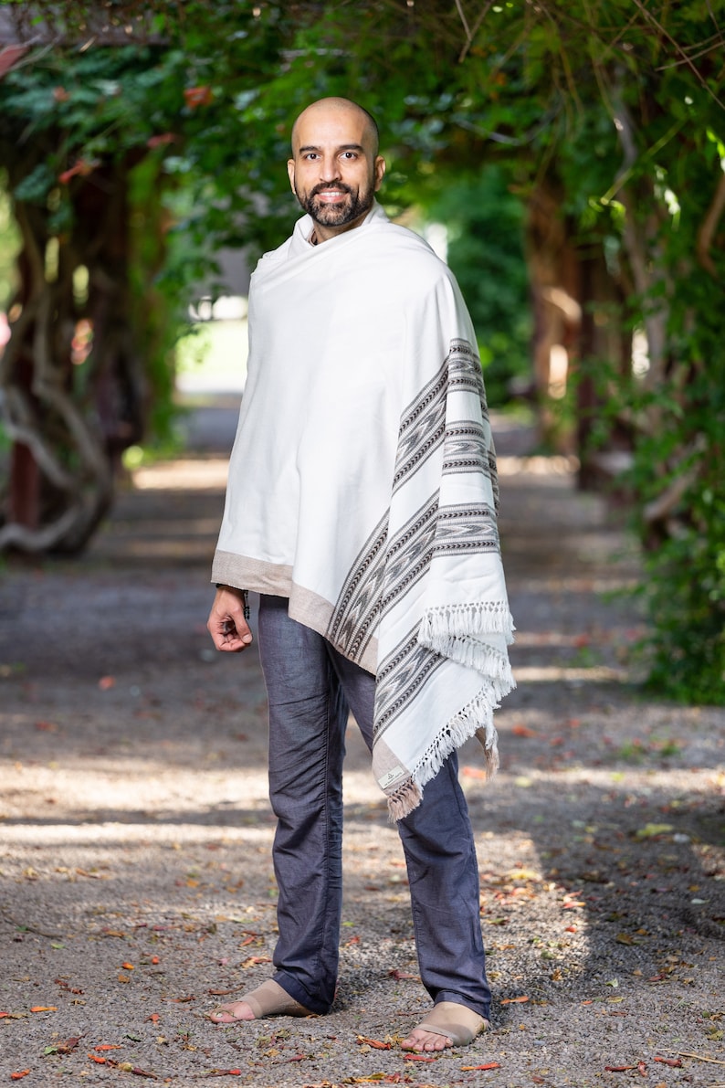 Puede incluir: Un hombre con un chal blanco con un patr&oacute;n geom&eacute;trico negro y marr&oacute;n. El chal tiene flecos en el borde inferior. El hombre lleva pantalones azules y est&aacute; de pie en un camino de grava.