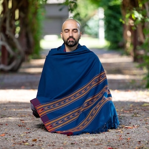 Puede incluir: Un hombre sentado con las piernas cruzadas en un camino, lleva una chal azul con rayas naranjas y rojas y borlas. Tiene los ojos cerrados y barba.