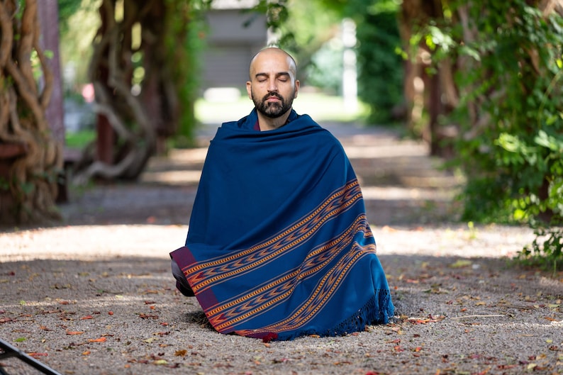 Puede incluir: Un hombre sentado con las piernas cruzadas en un camino, lleva una chal azul con rayas naranjas y rojas y borlas. Tiene los ojos cerrados y barba.