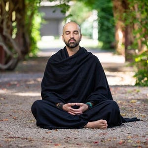 May include: A person with a shaved head and a beard sits in a cross-legged meditation pose. They are wearing a solid black, loose-fitting robe that drapes over their shoulders and body. Beaded bracelets are visible on both wrists.