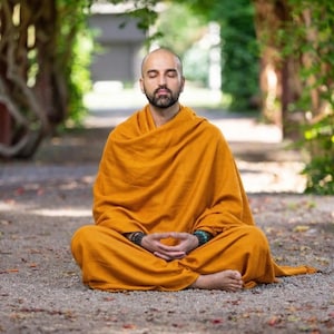 Puede incluir: Una persona en posición de meditación sentada, vestida con una túnica naranja, con los ojos cerrados. La persona está sentada sobre una superficie de grava, rodeada de árboles y vegetación. La túnica está drapeada sobre los hombros y las piernas.
