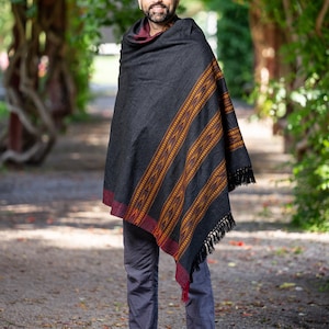 Puede incluir: Un hombre con un chal negro con un patr&oacute;n geom&eacute;trico rojo y naranja. El chal tiene flecos en los bordes. Lleva pantalones vaqueros azules y est&aacute; descalzo. Est&aacute; de pie en un camino de grava.