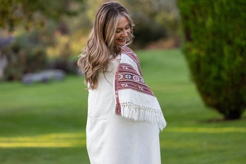 K&ouml;nnte beinhalten: Eine Frau in einem wei&szlig;en Kleid mit einem roten und wei&szlig;en bestickten Schal. Der Schal hat Fransen und ein geometrisches Muster. Sie steht auf einer gr&uuml;nen Wiese.