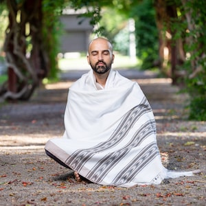Puede incluir: Un hombre se sienta con las piernas cruzadas en una postura meditativa, con un chal blanco con patrones geom&eacute;tricos negros y grises. Tiene los ojos cerrados y una expresi&oacute;n de paz en su rostro. El fondo es una imagen borrosa de un arco verde frondoso.