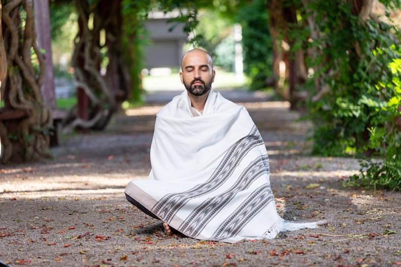 Puede incluir: Un hombre se sienta con las piernas cruzadas en una postura meditativa, con un chal blanco con patrones geom&eacute;tricos negros y grises. Tiene los ojos cerrados y una expresi&oacute;n de paz en su rostro. El fondo es una imagen borrosa de un arco verde frondoso.