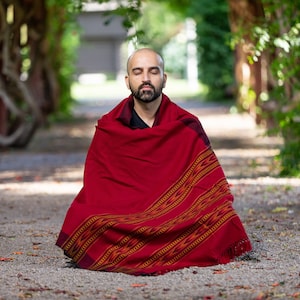 Puede incluir: Un hombre se sienta con las piernas cruzadas en un camino de un jard&iacute;n, meditando con los ojos cerrados. Lleva una chalina roja y negra con estampado.