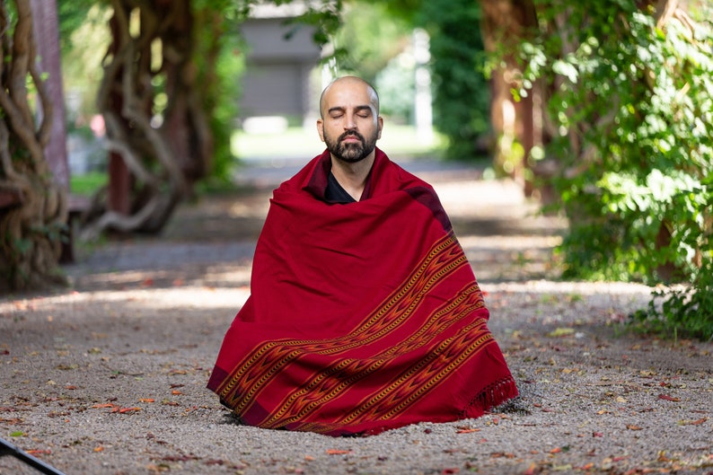 Puede incluir: Un hombre se sienta con las piernas cruzadas en un camino de un jard&iacute;n, meditando con los ojos cerrados. Lleva una chalina roja y negra con estampado.