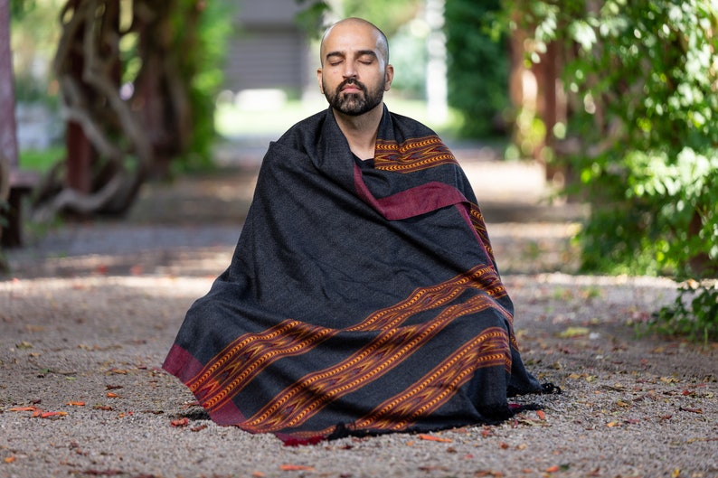 Puede incluir: Un hombre est&aacute; sentado con las piernas cruzadas en un camino, meditando con los ojos cerrados. Lleva una chal de color negro con un patr&oacute;n rojo.