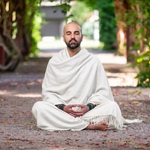 Puede incluir: Una persona en pose meditativa, sentada al aire libre con los ojos cerrados. Está envuelta en un chal blanco, con las manos en un mudra. El entorno es un sendero arbolado, con luz moteada y un ambiente sereno.