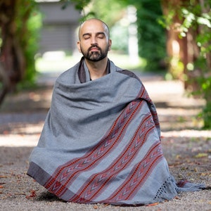Puede incluir: Un hombre est&aacute; sentado con las piernas cruzadas en el suelo, envuelto en una manta de color gris y rojo con patrones. Tiene los ojos cerrados y parece estar meditando.