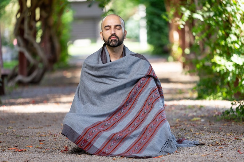 Puede incluir: Un hombre est&aacute; sentado con las piernas cruzadas en el suelo, envuelto en una manta de color gris y rojo con patrones. Tiene los ojos cerrados y parece estar meditando.