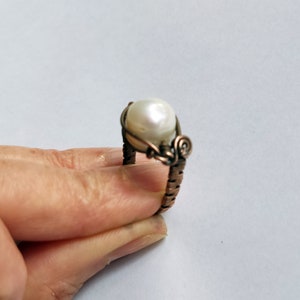 May include: A white pearl ring with a copper wire design. The ring is wrapped in copper wire and has a swirl design on the side.