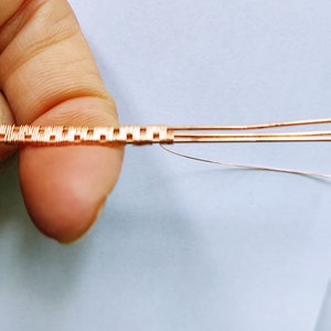 May include: Close-up of a copper wire woven into a square pattern. The wire is held in a hand, and the background is a light blue surface.
