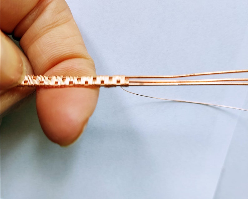 May include: Close-up of a copper wire woven into a square pattern. The wire is held in a hand, and the background is a light blue surface.