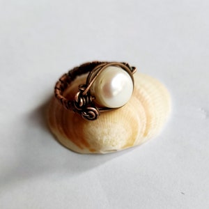 May include: A copper wire ring with a white pearl wrapped in the wire. The ring is sitting on a seashell.