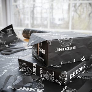 May include: Two black gift boxes wrapped in paper with the word "BECOME" in white. The wrapping paper has a repeating pattern of the word "BECOME" and a monkey graphic. A pair of scissors and a roll of wrapping paper are also visible.