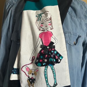 May include: A long scarf with a whimsical design. The scarf features a hand-drawn illustration of a girl walking a dog, with a pink top and a black skirt. The scarf has black, teal, and white panels with a black velvet backing.