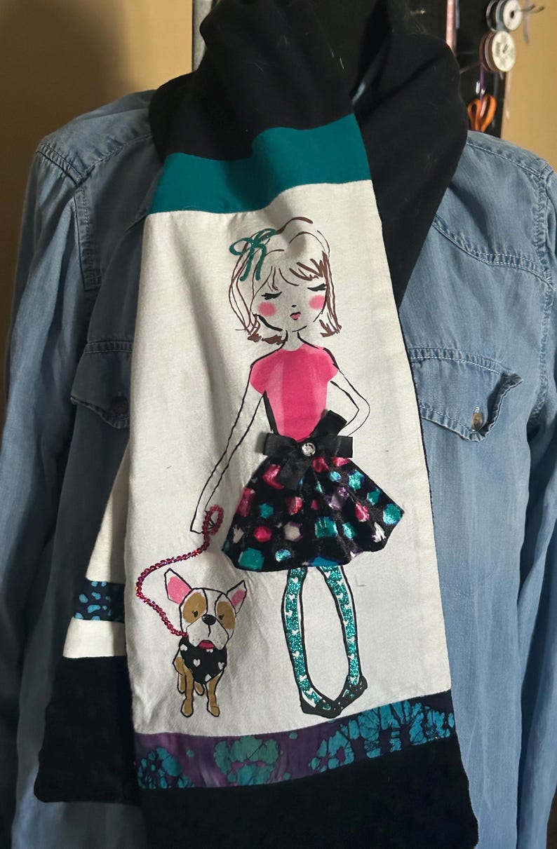 May include: A long scarf with a whimsical design. The scarf features a hand-drawn illustration of a girl walking a dog, with a pink top and a black skirt. The scarf has black, teal, and white panels with a black velvet backing.