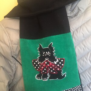 May include: Black scarf with a green panel featuring a black dog with a red and white polka dot bow. The scarf has a black and white polka dot border and is made of a soft material.