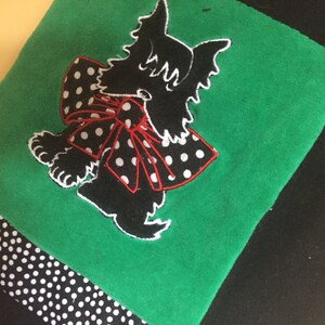 May include: A green fabric item featuring a black silhouette of a dog with a red bow with white polka dots. The bottom edge has a black and white polka dot pattern. The dog has white outlines.