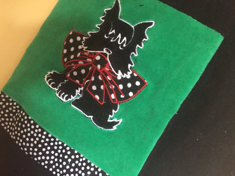 May include: A green fabric item featuring a black silhouette of a dog with a red bow with white polka dots. The bottom edge has a black and white polka dot pattern. The dog has white outlines.