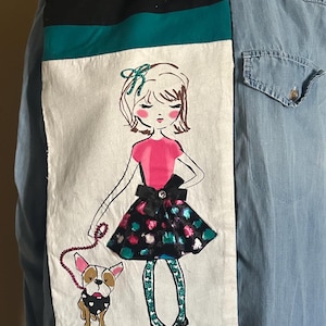 May include: A decorative scarf featuring a hand-drawn illustration of a girl in a pink top and a patterned skirt, walking a bulldog. The scarf has a black, teal, and white color scheme with a purple and teal patterned border.