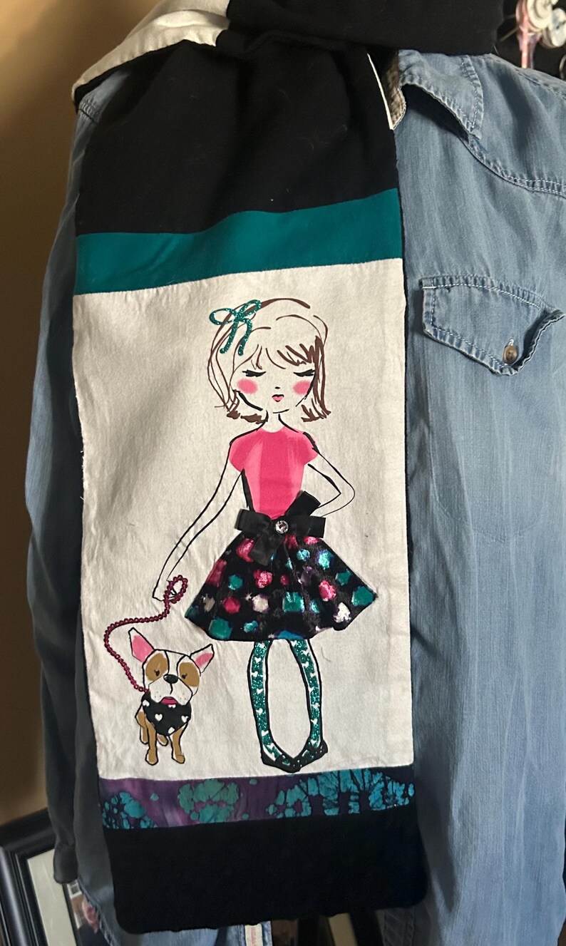 May include: A decorative scarf featuring a hand-drawn illustration of a girl in a pink top and a patterned skirt, walking a bulldog. The scarf has a black, teal, and white color scheme with a purple and teal patterned border.