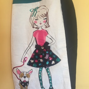 May include: A decorative hand towel featuring a whimsical illustration of a girl and her dog. The girl wears a pink top and a sequined skirt. The dog is a French Bulldog. The towel has a teal and black border.