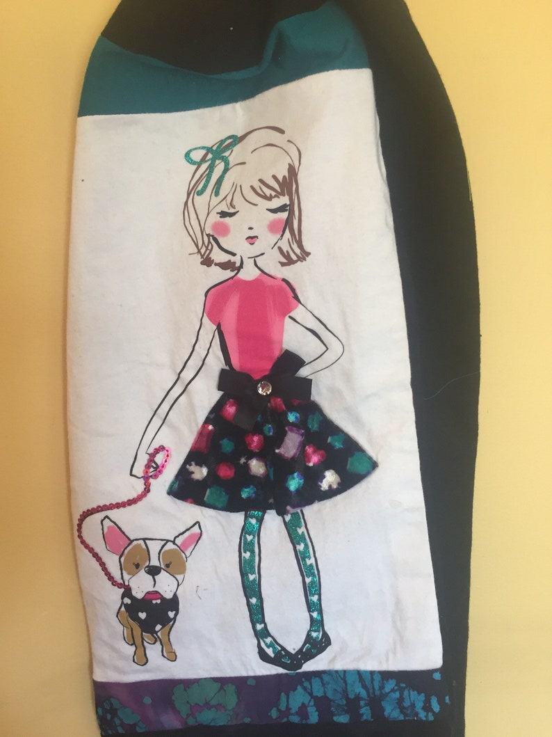 May include: A decorative hand towel featuring a whimsical illustration of a girl and her dog. The girl wears a pink top and a sequined skirt. The dog is a French Bulldog. The towel has a teal and black border.