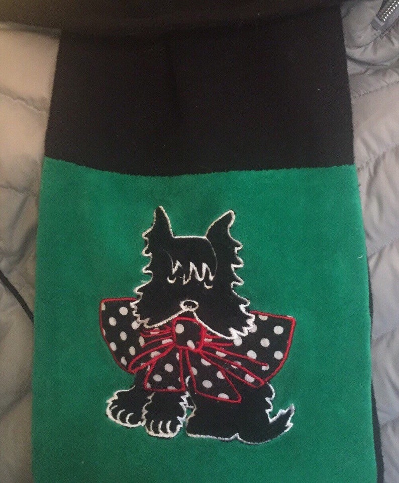 May include: A black and green scarf featuring an embroidered black Scottish Terrier dog with a white polka dot bow and red trim. The top portion of the scarf is black.