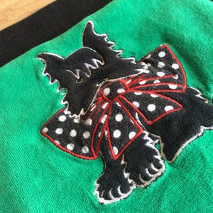 May include: A green towel with a black terrier dog applique. The dog has a white outline and a red and white polka dot bow. The towel is a solid green color.