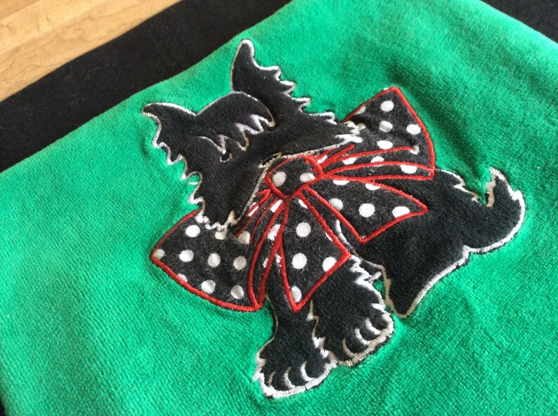 May include: A green towel with a black terrier dog applique. The dog has a white outline and a red and white polka dot bow. The towel is a solid green color.