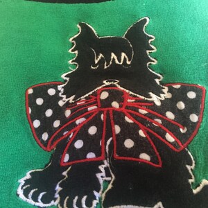 May include: A green textile with a black dog applique. The dog has a white outline, a black body, and a bow with white polka dots and a red outline. The dog's ears are pointed and the bow is centered.