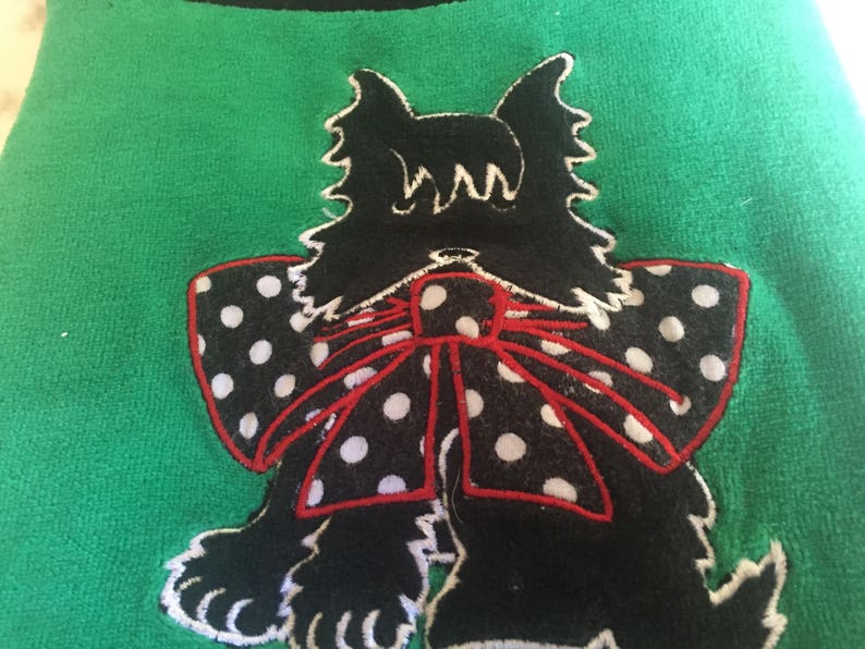 May include: A green textile with a black dog applique. The dog has a white outline, a black body, and a bow with white polka dots and a red outline. The dog's ears are pointed and the bow is centered.