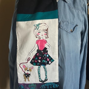 May include: A long, rectangular scarf with a black top and bottom, a teal stripe, and a white center panel. The center panel features a drawing of a girl in a pink top and a patterned skirt, walking a dog. The scarf is displayed on a denim shirt.
