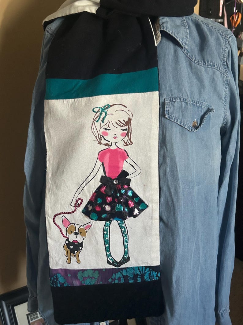 May include: A long, rectangular scarf with a black top and bottom, a teal stripe, and a white center panel. The center panel features a drawing of a girl in a pink top and a patterned skirt, walking a dog. The scarf is displayed on a denim shirt.