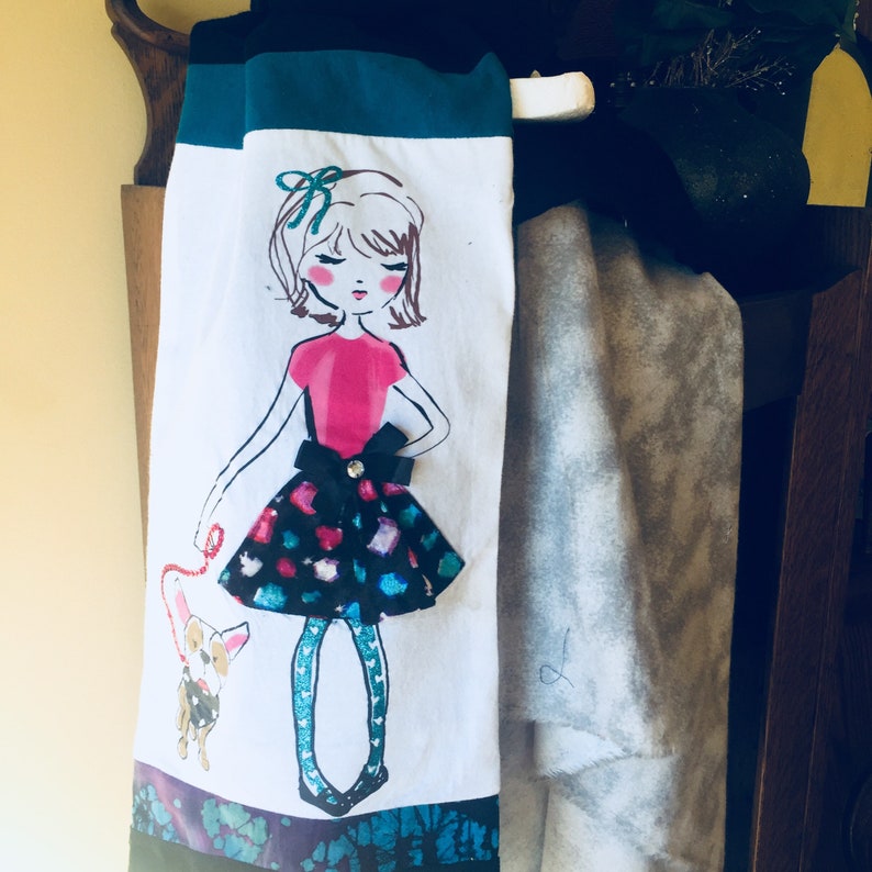May include: White decorative towel featuring a hand-drawn illustration of a girl in a pink top and a patterned skirt, holding a leash attached to a small dog. The towel has teal and black accents.