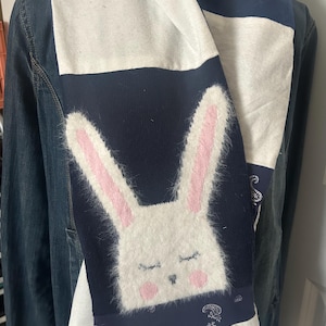 May include: A navy blue and white scarf featuring a large, fluffy white bunny face with pink inner ears and closed eyes. The scarf is draped over a denim jacket, showcasing its design and color scheme.