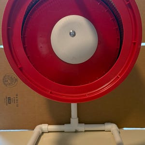 May include: A red, circular object with a white center and a mesh screen, supported by a white PVC pipe frame. The frame is rectangular and has a square base. The object appears to be a sifter or a similar tool.