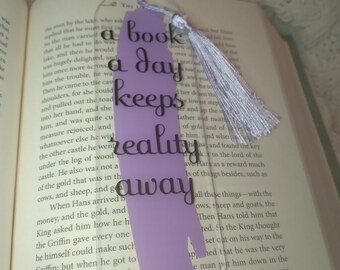 May include: A lavender bookmark with a tassel. The bookmark has the text "A book a day keeps reality away" in black lettering.