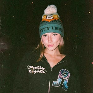May include: A person wearing a teal and yellow beanie with the text "Pretty Lights" and a black zip-up hoodie. The hoodie has the text "Pretty Lights" on the chest and a colorful graphic on the front.