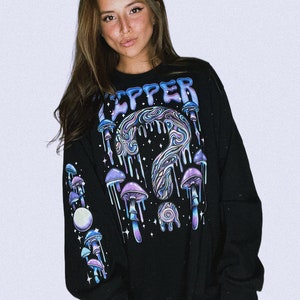 Puede incluir: Camiseta negra de manga larga con un gráfico psicodélico de hongos y el texto "Tripper" en una fuente que gotea. El gráfico presenta un diseño en espiral con estrellas y una luna.